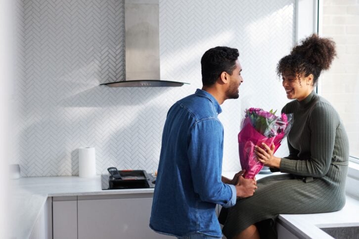Romantic Couple At Home With Man Surprising Woman With Bunch Of Flowers Celebrating Valentines Day
