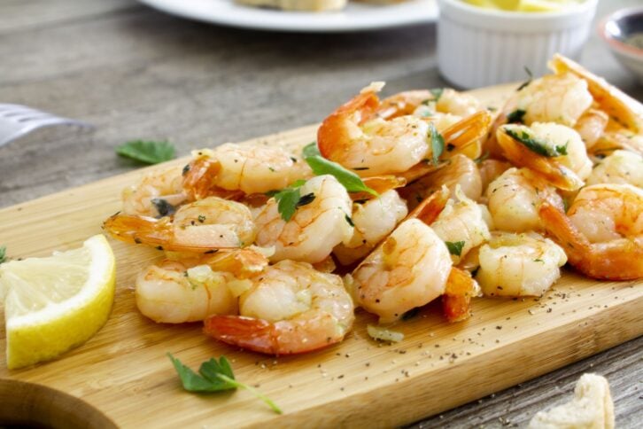 Panfried shrimp with garlic and herbs served on a white plate with lemon and fresh crusty bread.Shot on a wooden chopping board on a rustic wooden table. Close up.