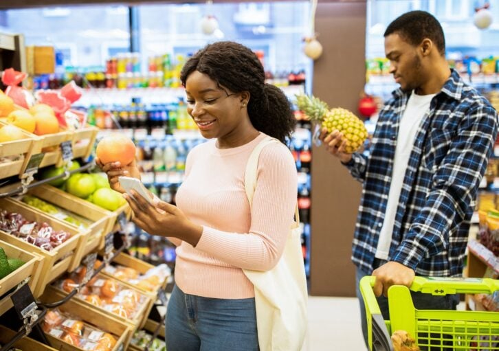 Lovely black woman and her boyfriend shopping for fruits, checking grocery list on mobile phone at supermarket. African American couple choosing healthy food at produce section of mall
