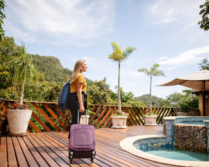 Smiling young woman with a wheeled suitcase arriving at a tourist resort in the tropics