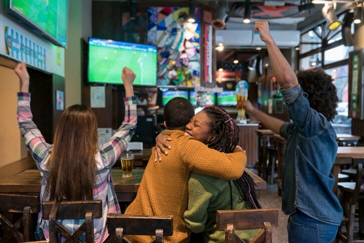 Back view of diverse friends celebrating Argentina's triumph on a football match at the bar hugging while enjoying beer