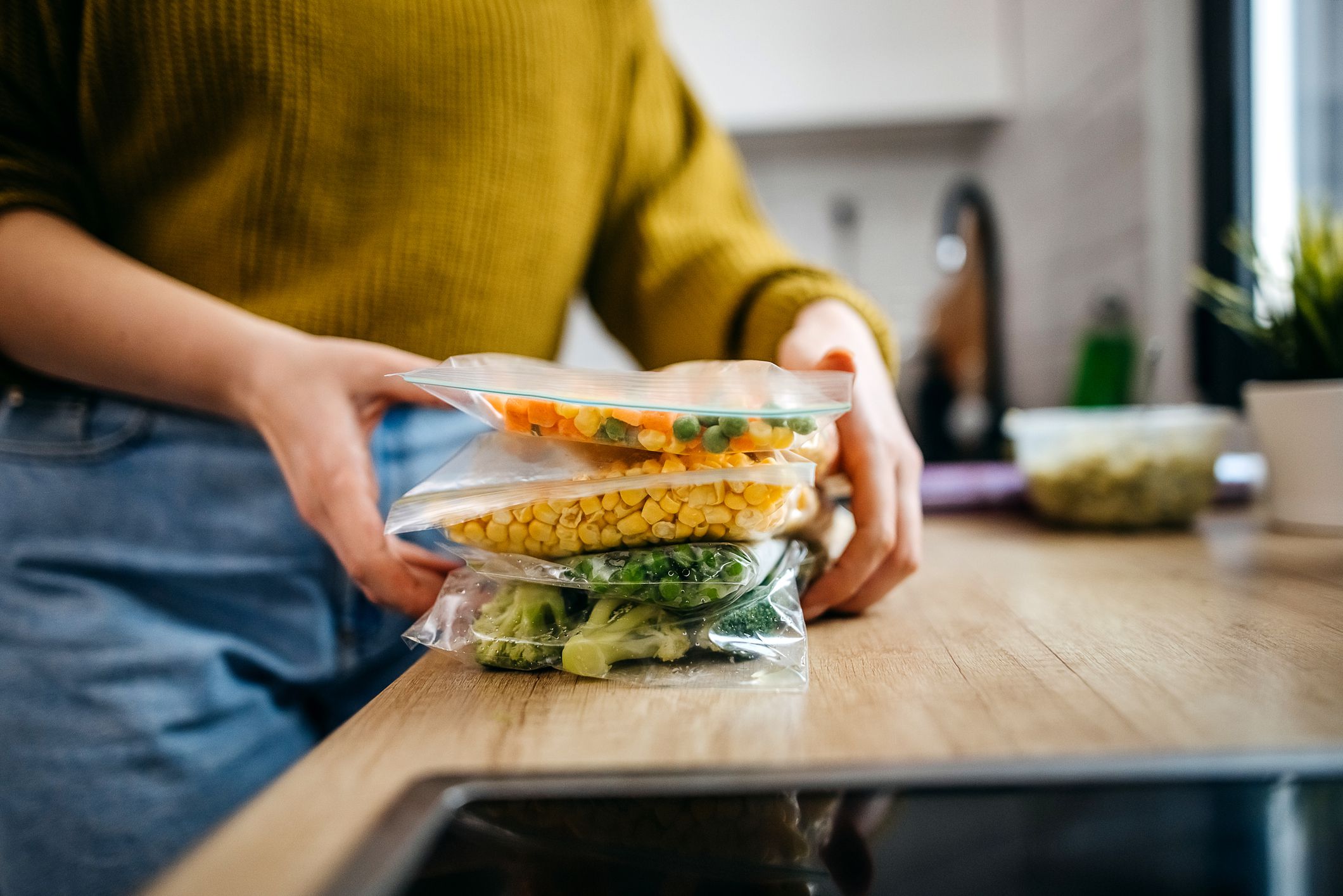 A person wearing a green sweater holds three stacked ziplock bags filled with chopped frozen vegetables, including carrots, corn, and broccoli, on a kitchen counter.