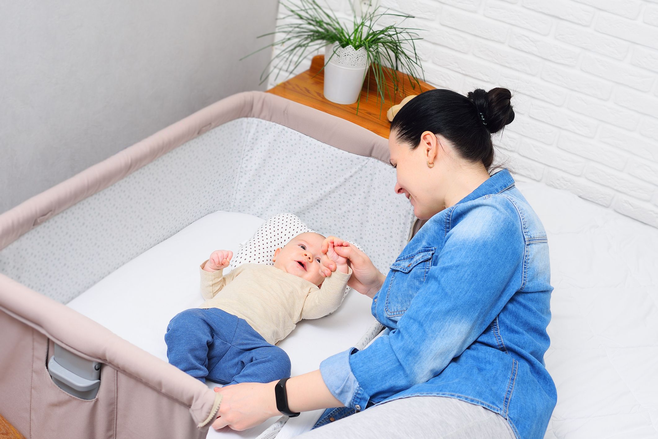 a mother plays with a baby who is lying in a side-bed for a newborn or in a modern playpen
