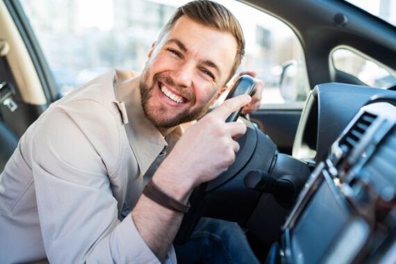 A smiling man in a light-colored jacket sitting in the driver’s seat of a car and holding the steering wheel.