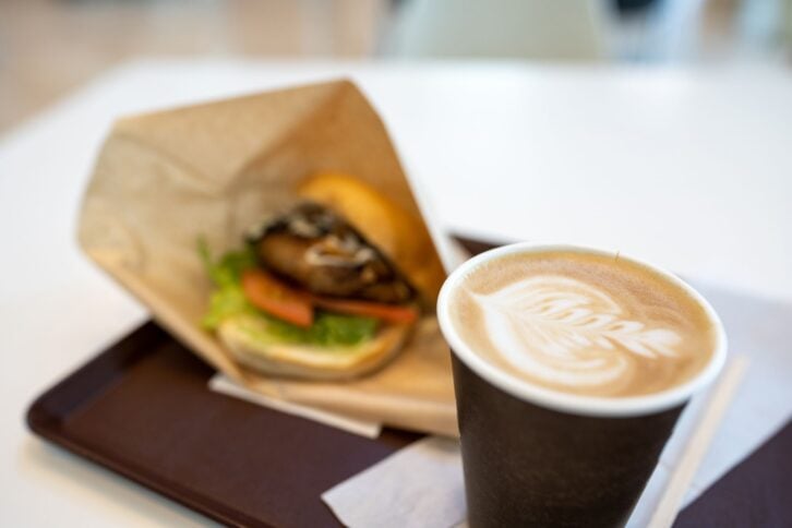 Cappuccino with latte art and fresh burger blurred in the background on a tray