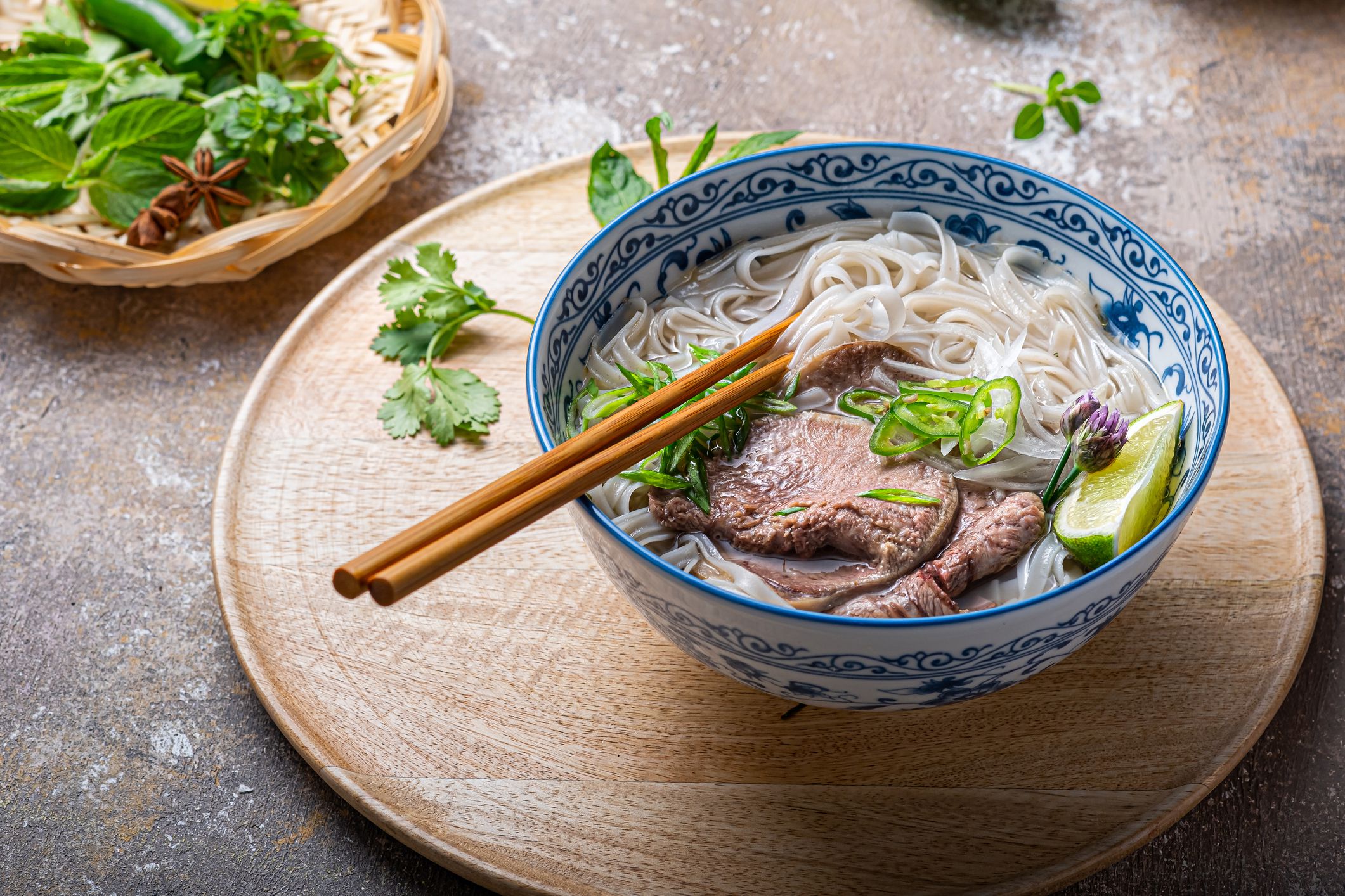 Close view of pho bo in traditional bowl, garnished with basil, mint, lime, on concrete background.