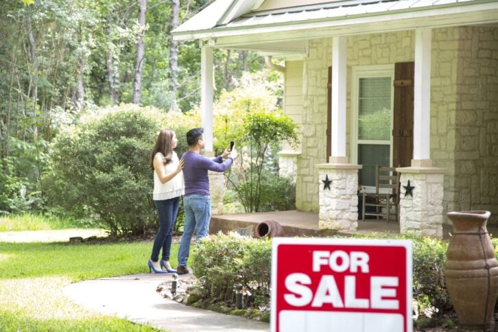 Mid adult Asian couple are considering purchasing a new home.  For sale sign in foreground.