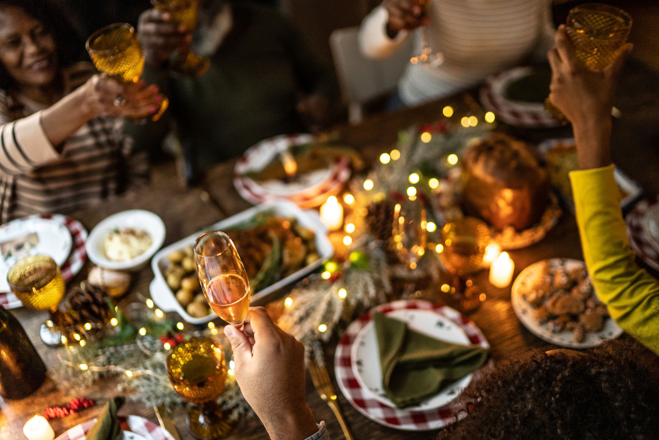 People gathered around a festive dinner table, raising glasses in a toast. The table is decorated with candles, lights, holiday greenery, and various dishes, creating a warm, celebratory atmosphere.
