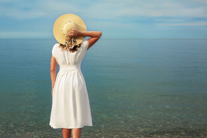 A young girl in a white dress stands on the seashore, looking into the distance. Holds the straw hat with his right hand