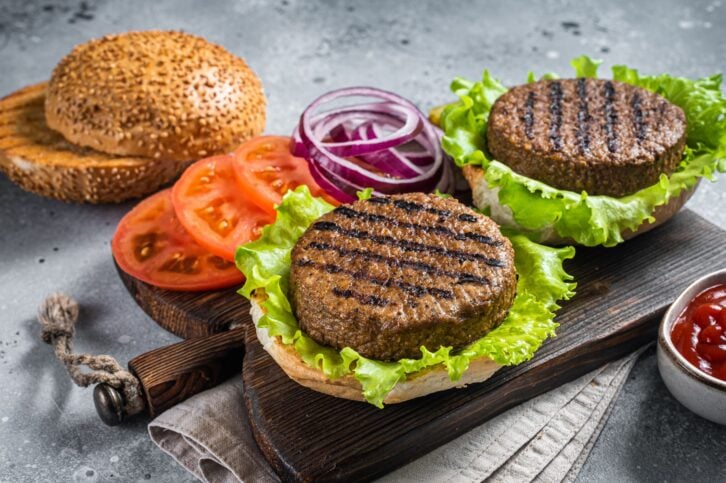 Plant based meatless burgers with vegan grilled pattie, tomato and onion on a wooden serving board. Gray background. Top view.
