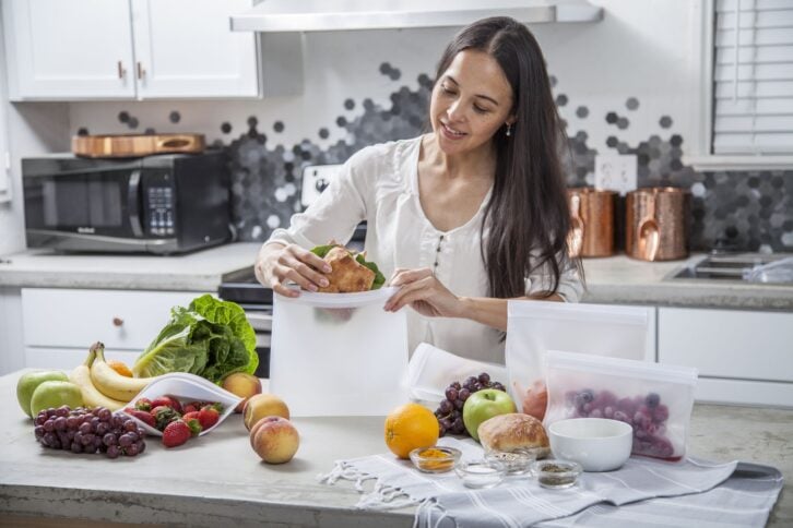 A young mother puts a sandwich into a clear food storage bag. She is in the kitchen with a concrete counter top surrounded by other fruits and vegetables that she is preparing and storing.