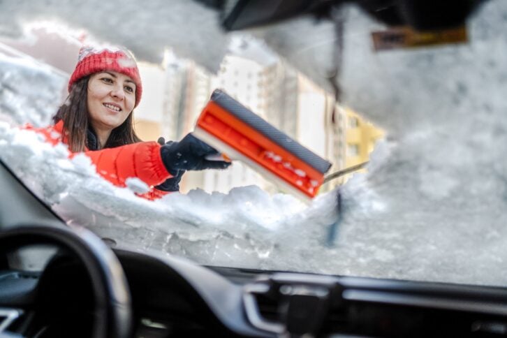 Millennial Woman in winter jacket scraping ice and snow from car windows
