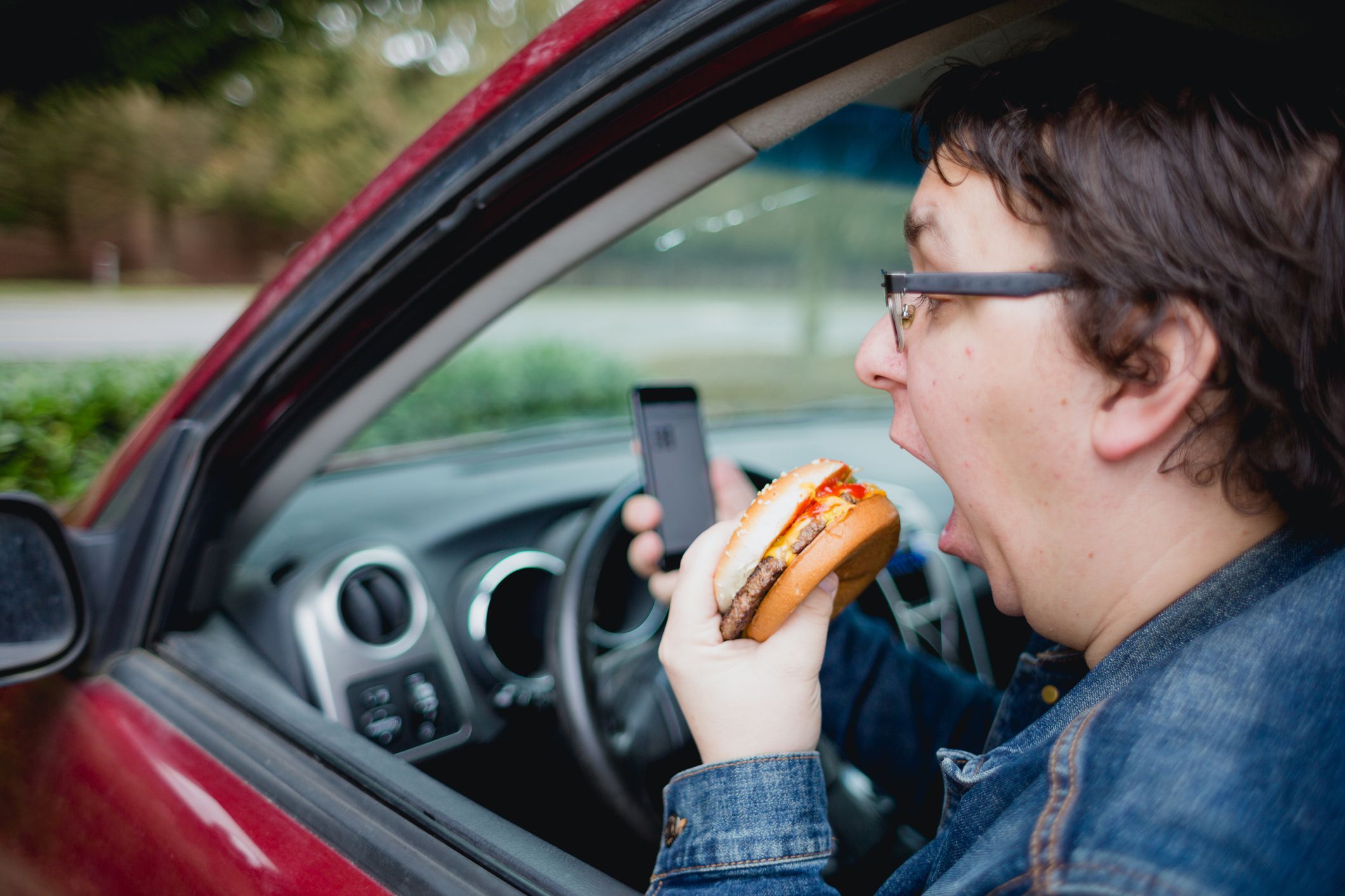 Man distracted by his cell phone while eating a hamburger for lunch in his car. Car is parked and he is not driving while using phone.