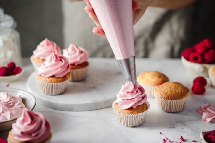 Close-up of a woman decorating muffins with pink whipped creamon the kitchen counter.  Female hands making raspberry cupcakes in kitchen.