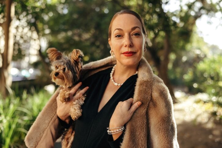 Elegant woman looking at the camera while holding her puppy outdoors. High class woman standing alone in a park during the day. Female pet owner going for a walk with her dog.
