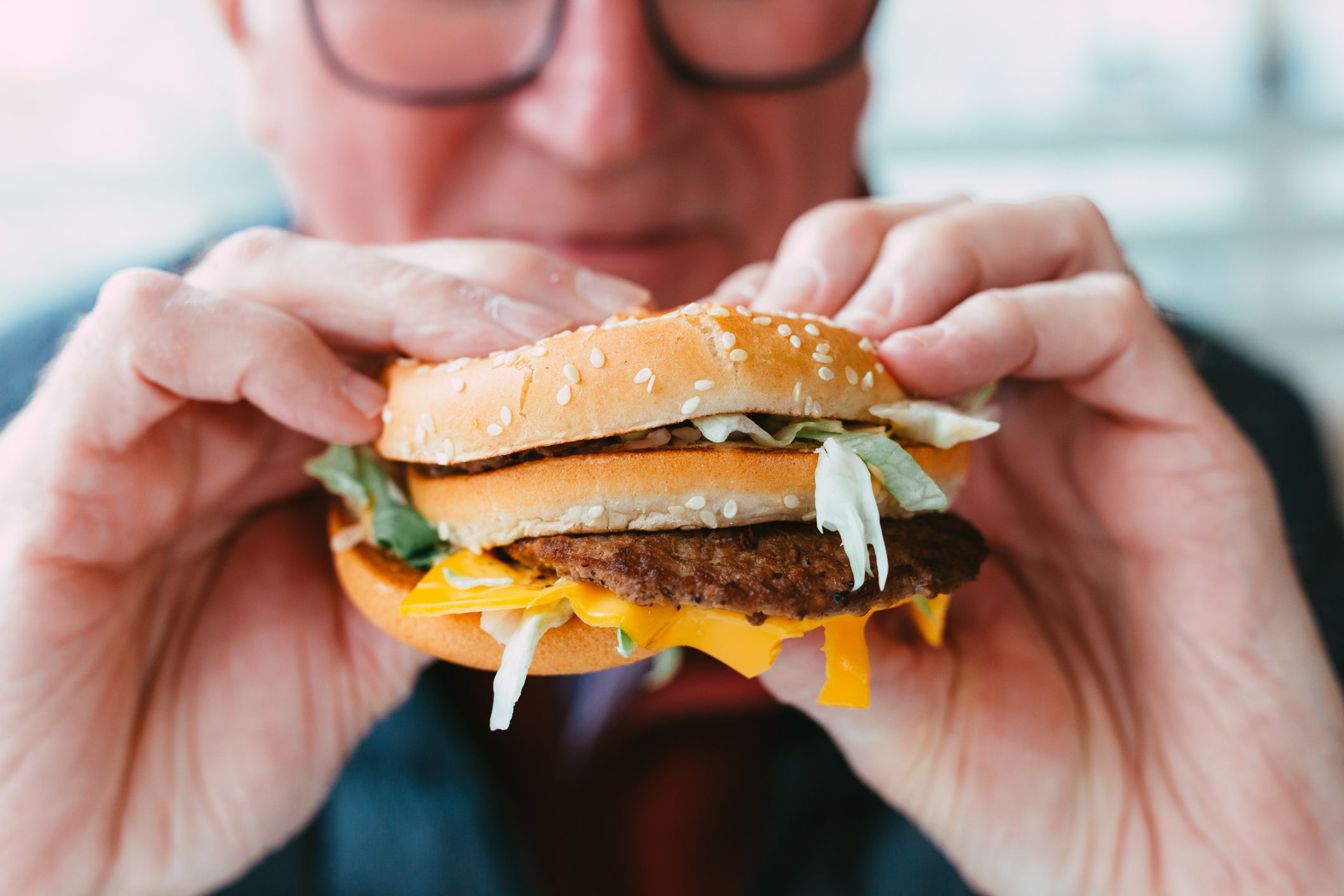 Close up color image depicting a senior man wearing spectacles eating a freshly cooked burger in a fast food restaurant. The burger is messy, with lettuce and cheese hanging out of the bun. Room for copy space.