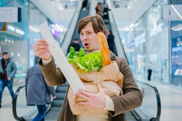 Man in a coat holds a paper check and a bag with a baguette and lettuce in a shopping mall and is shocked by the high prices of groceries.