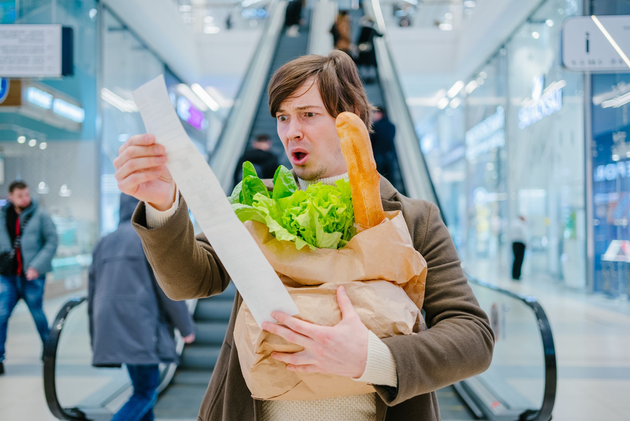 Man in a coat holds a paper check and a bag with a baguette and lettuce in a shopping mall and is shocked by the high prices of groceries.