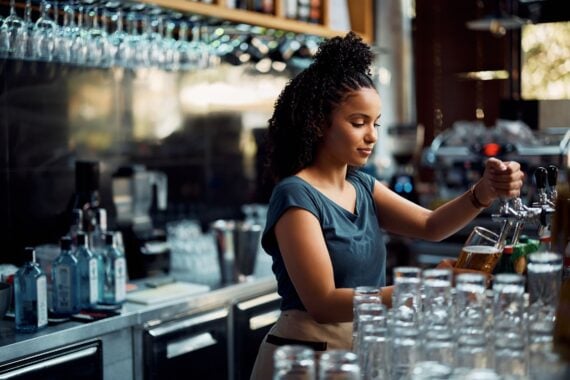 African American female barista pouring beer from a beer tap while working in pub.