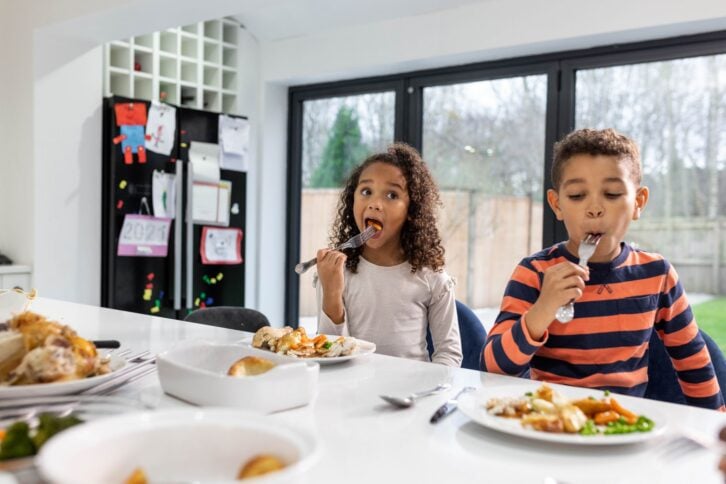 Two children placing food into their mouths looking away from camera. They are both sitting next to a kitchen worktop.