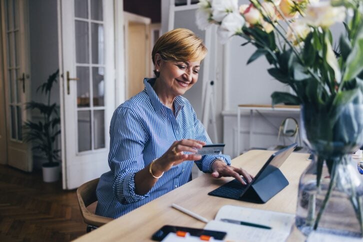 A smiling Caucasian female entrepreneur holding her credit card and typing on her tablet while sitting at the desk.