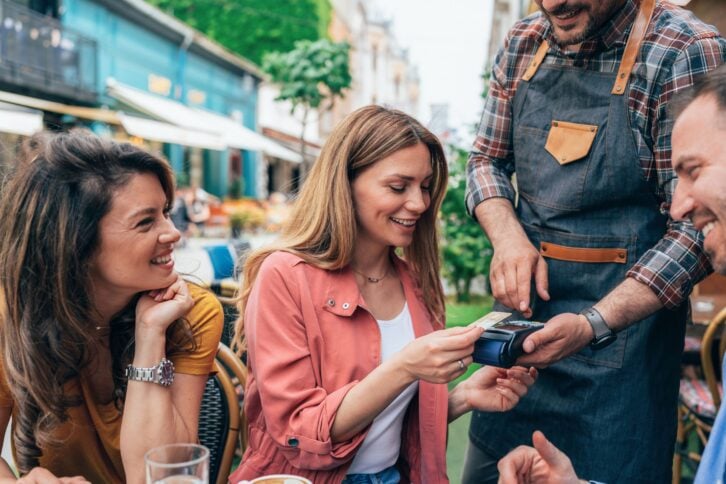 Young woman paying contactless using credit card in sidewalk cafe, with friends. Group of young people sitting in outdoor cafe, paying the bill
