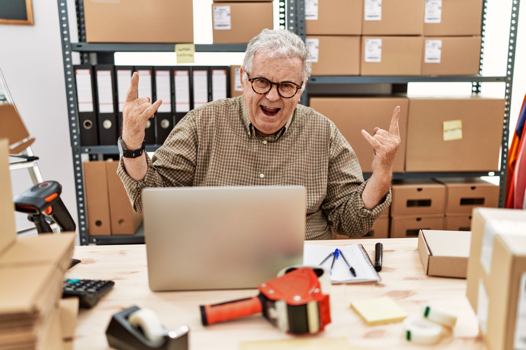 Senior caucasian man working at small business ecommerce with laptop shouting with crazy expression doing rock symbol with hands up. music star. heavy concept.