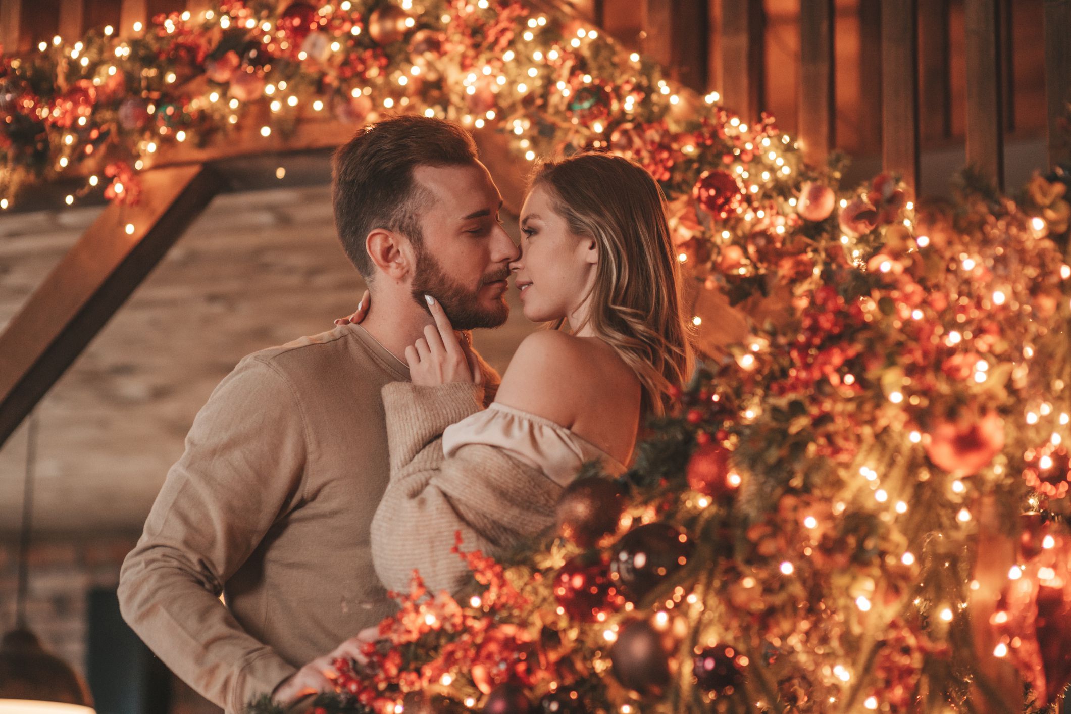 Portrait of young lovely couple hugging indoor eve 25 December. Lovers laughing hugs kisses waiting xmas at home. Celebrating new year garlands lights noel in elegant knitwear outfit tenderness