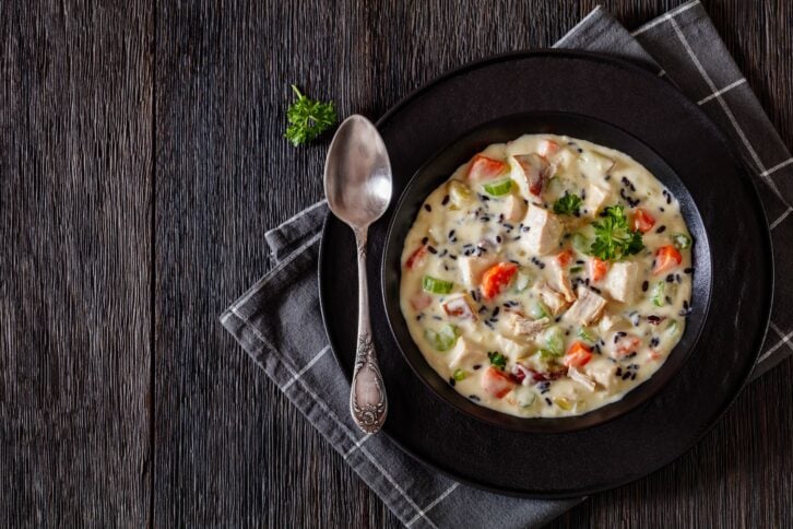 Turkey Wild Rice Soup with red potatoes. carrot, celery, spices and herbs in black bowl on dark wooden table with spoon, horizontal view from above, flat lay, free space