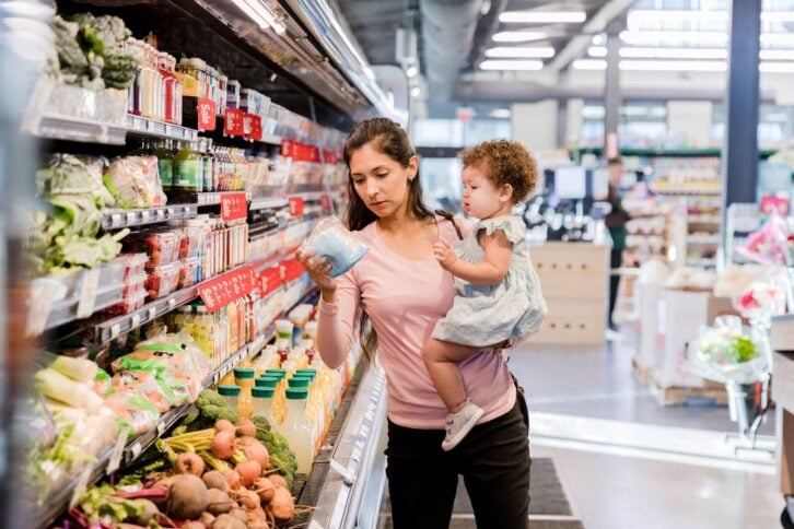 The young mother holds her daughter on her hip as she grocery shops for items she needs.