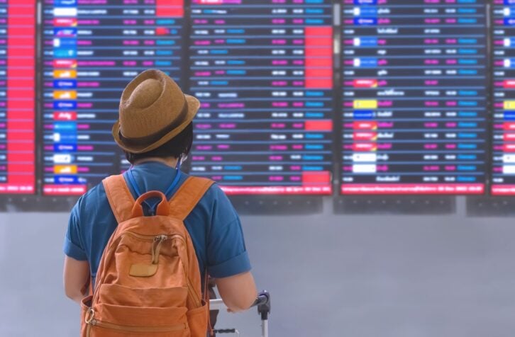 Rear view of female Tourist with backpack and luggage cart checking her flight on Digital Information Board in Terminal Airport