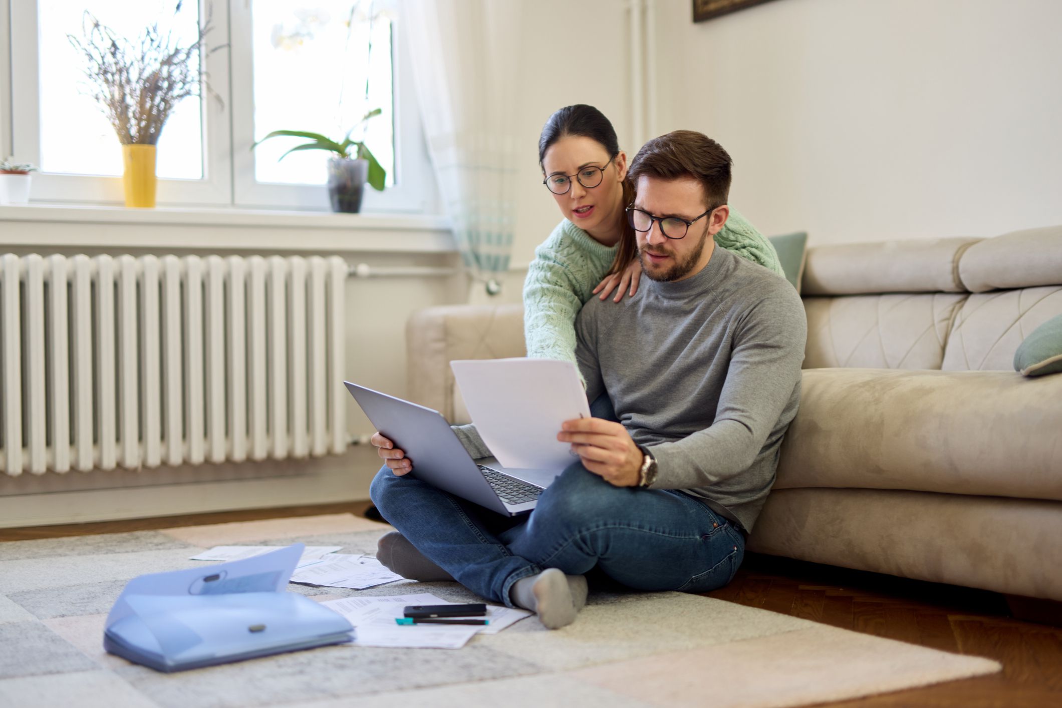 A couple sits on the floor at home, looking at papers and a laptop. The man holds documents while the woman leans over his shoulder. Bills and a calculator are spread around them, suggesting they are managing finances.