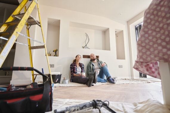A couple sits on the floor of a room under renovation, smiling and looking up. Around them are tools, paint cans, a ladder, and a toolbox. The walls are unfinished, and drop cloths cover the floor.