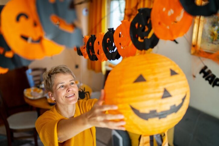 A woman is reaching up to hang a paper jack o' lantern while decorating a room ready for a halloween party