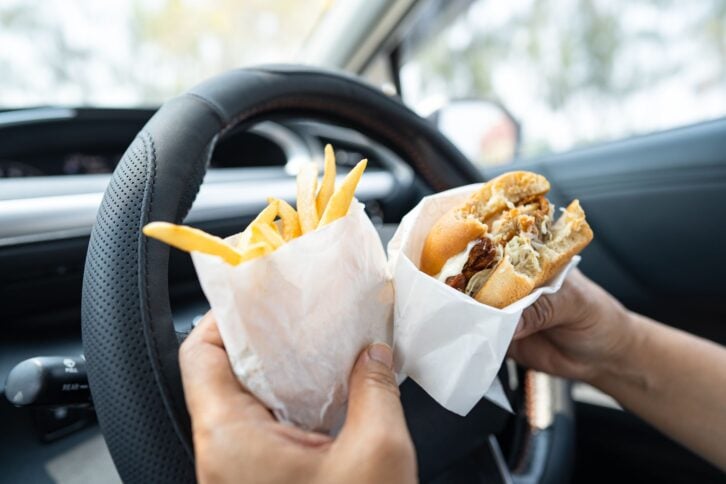 Asian lady holding hamburger and French fries to eat in car, dangerous and risk an accident.