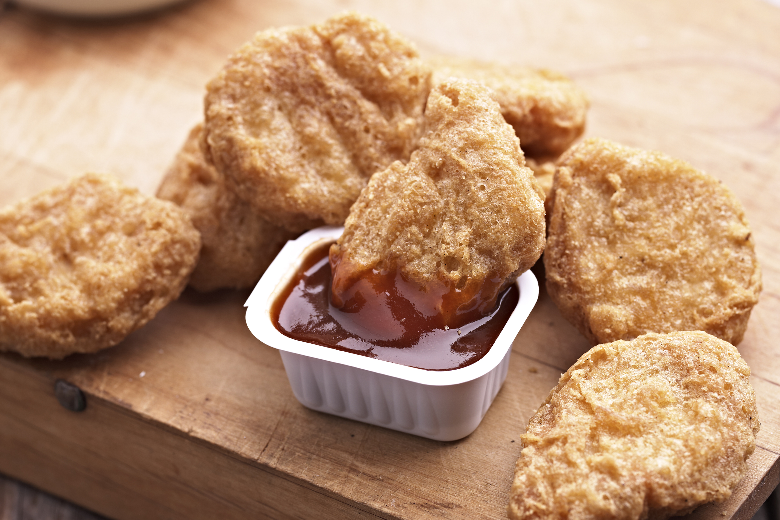 Several golden-brown chicken nuggets are arranged on a wooden surface, with one nugget being dipped into a small container of reddish-brown sauce.