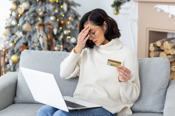 Sad young woman sitting at home on the sofa for the holidays near the Christmas tree, holding a credit card and holding her head worriedly.