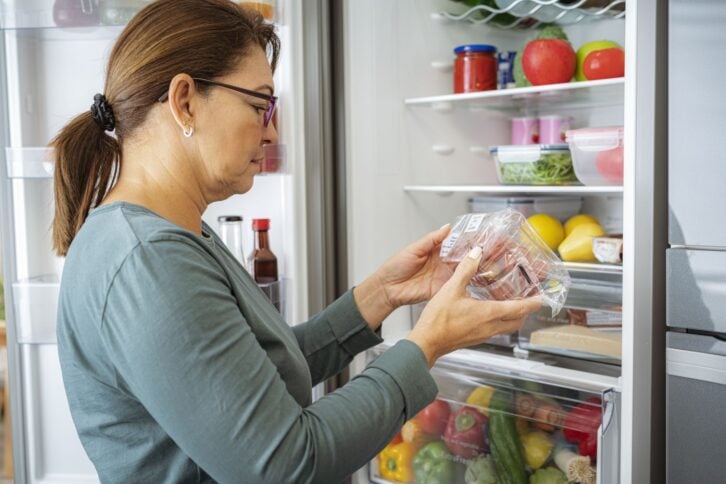 Woman by the fridge reading label of refrigerated food package