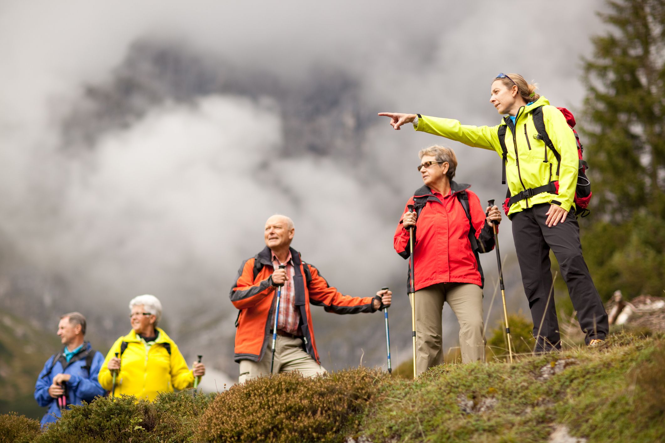 young female hiking guide showing senior group surrounding mountains, shallow focus, focus on young woman