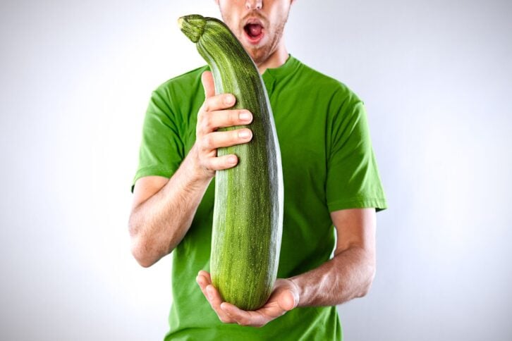A young man gasps at a massive farm grown organic zucchini with a green shirt to match.  Horizontal with copy space.