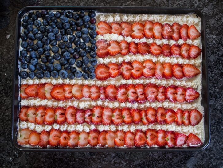 A patriotic dessert featuring a cake adorned with strawberries, blueberries, and cream, beautifully arranged to resemble the American flag.
