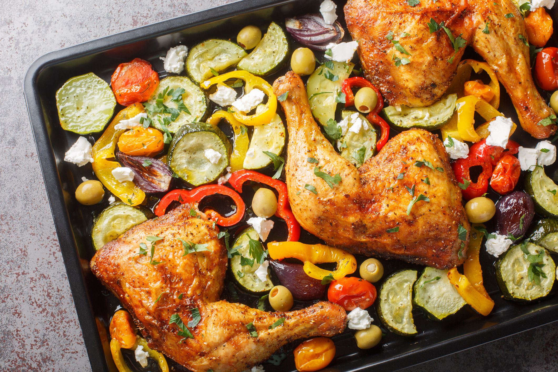 Greek style baked chicken quarters with zucchini, tomatoes, peppers, olives and feta cheese close-up on a baking sheet on the table. Horizontal top view from above
