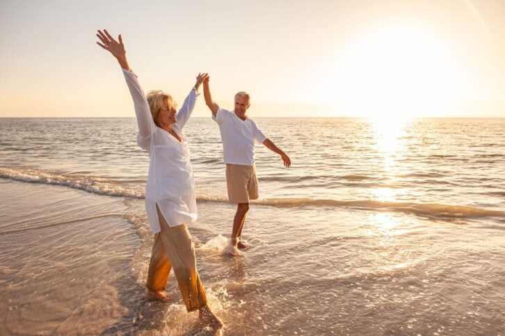 Happy senior man and woman old retired couple walking and holding hands on a beach at sunset