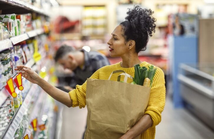A mature Hispanic woman, dressed in a yellow sweater, selects products in a grocery store, carrying a paper bag with fresh vegetables peeking out.