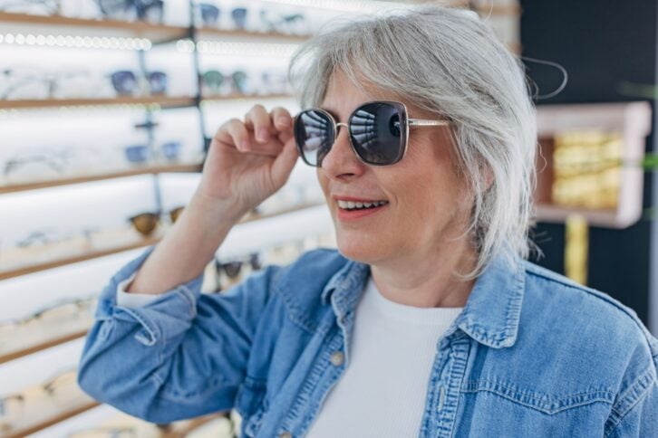 A woman with gray hair tries on dark sunglasses in an eyeglass store, adjusting the frames with a smile while standing in front of shelves filled with various eyeglasses.