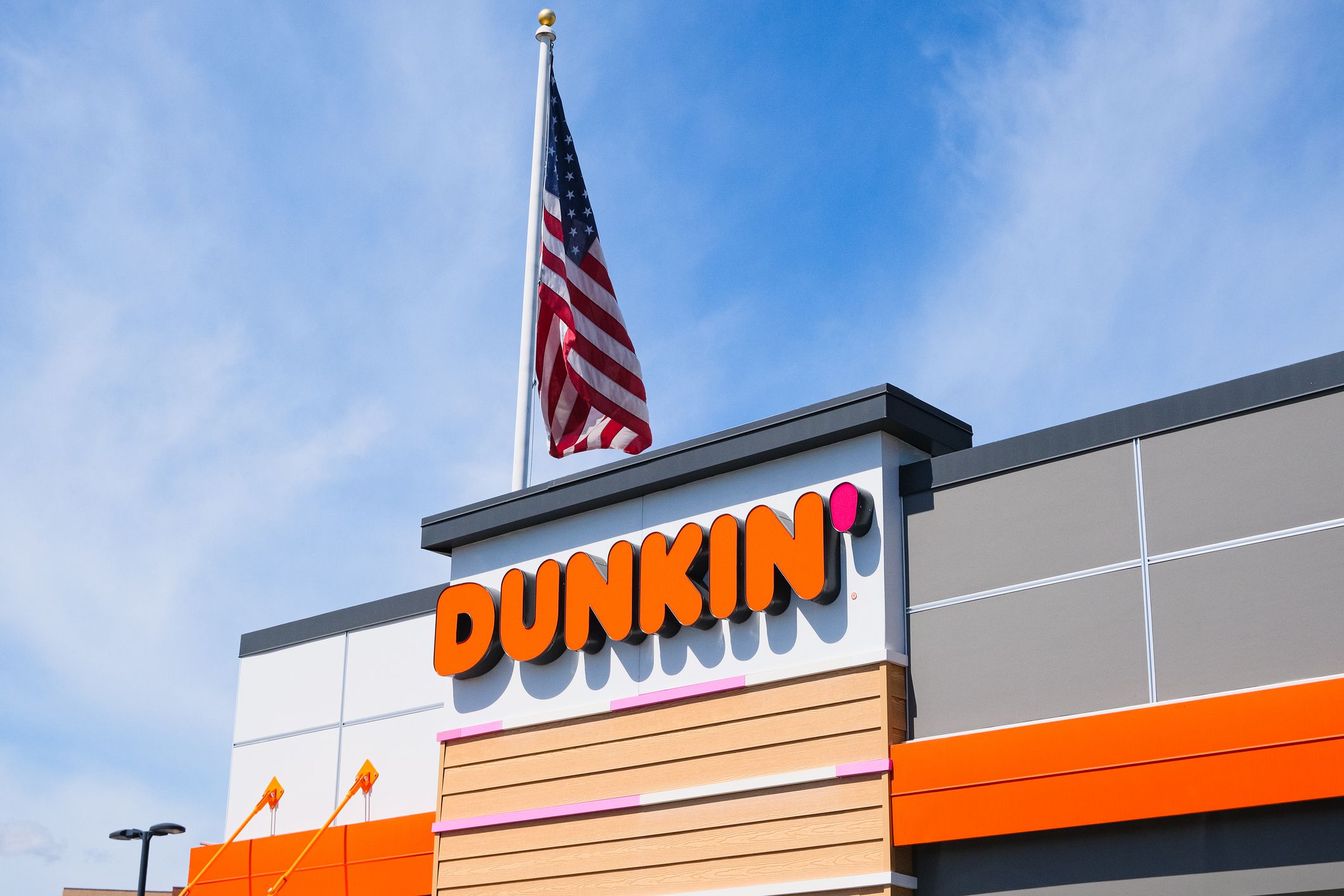 Massachusetts, USA - 19 April 2024. General view of the Dunkin Donuts logo sign on a cafe facade with American flag on flagpole on blue sky background