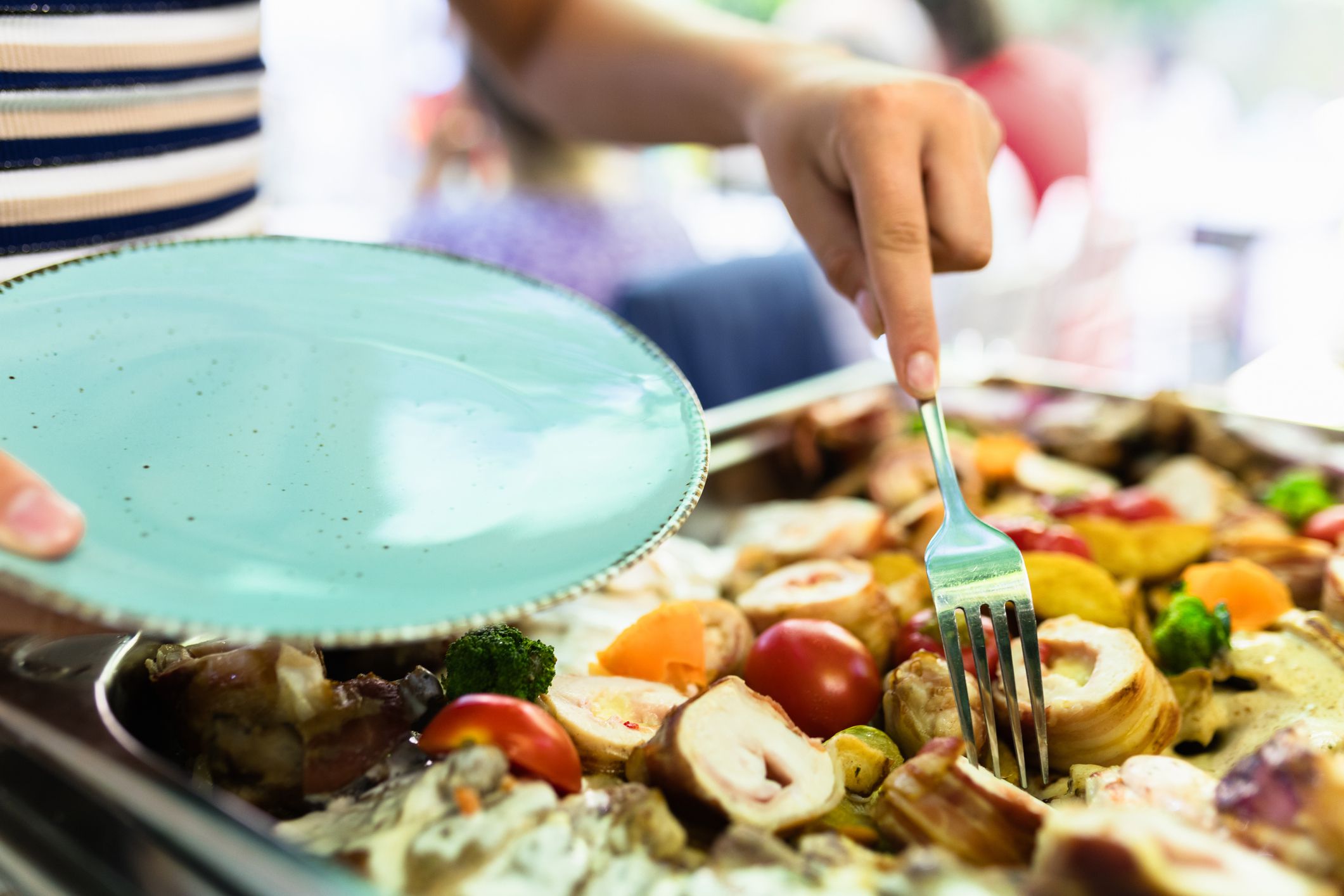 A person is plating a vibrant selection of roasted vegetables and meats at a cheerful brunch gathering outdoors, with a focus on sharing and enjoyment among friends.