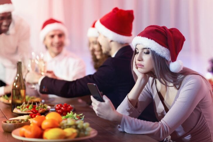 Boring New Year Party. Sad Girl Texting On Phone During Dreadful Christmas Dinner With Friends Sitting At Celebratory Table Indoor