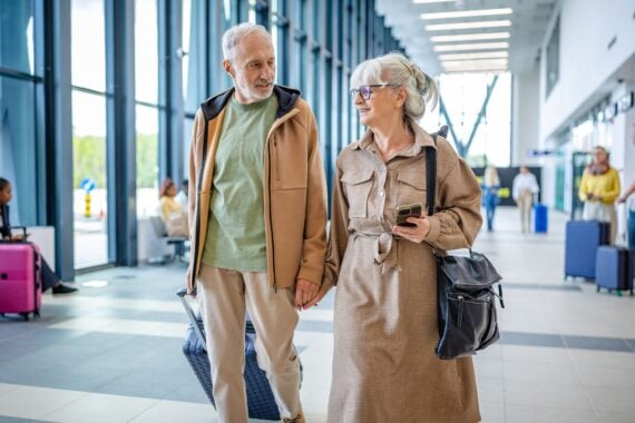 An elderly couple walks hand in hand through a modern airport terminal, navigating toward their flight with luggage in tow, displaying companionship and travel preparations.