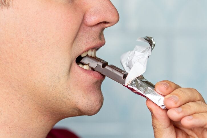 Portrait of young handsome man eating chocolate against blue background. A man's mouth in close-up with white teeth biting dark chocolate.